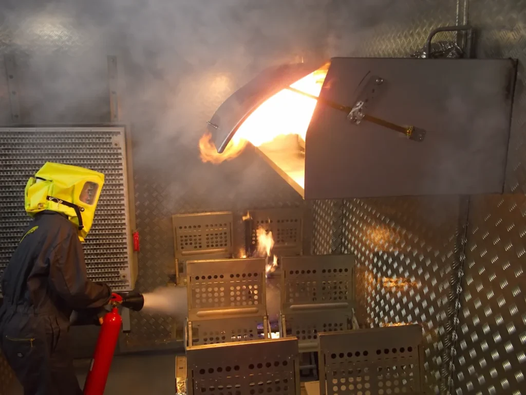 A trainee wearing a yellow protective breathing hood uses an extinguisher in a smoky fire-training cabin.